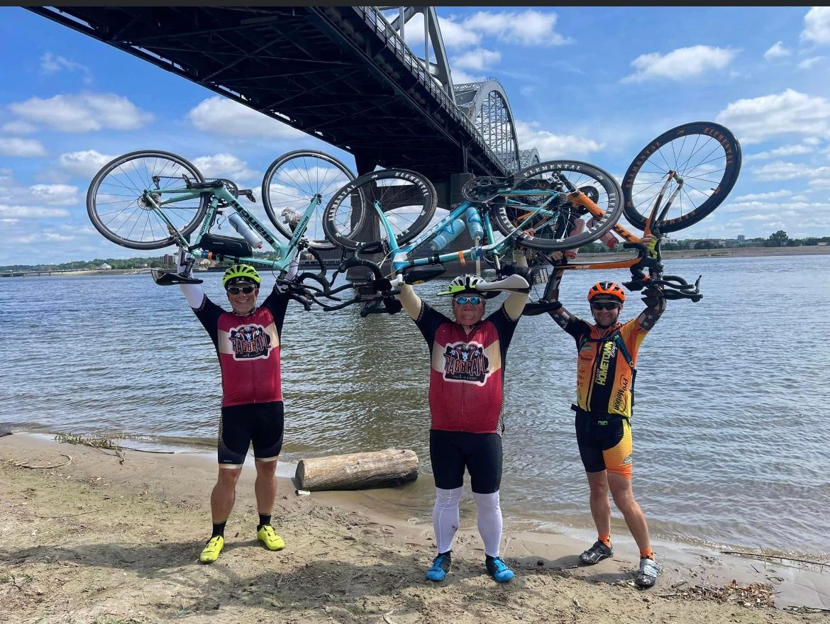 Three riders holding bikes overhead by a river bridge